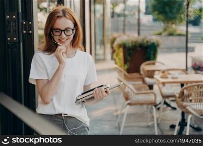 Happy beautiful redhead young woman makes distant call uses earphones with microphone carries modern gadgets poses outdoor at street against cafe blurred background. People and lifestyle concept. Beautiful redhead young woman makes distant call uses earphones with microphone