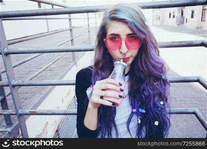 Happy beautiful teen with pink sunglasses drinks and enjoys a pink beverage sitting on urban ground