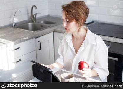 Happy beautiful woman with cup of coffee or tea using laptop in quarantine lockdown in the kitchen at home in the white shirt. Social distancing Self Isolation. Happy beautiful woman with cup of coffee or tea using laptop in quarantine lockdown in the kitchen in the white shirt.
