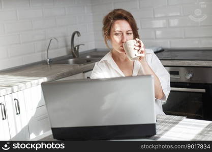 Happy beautiful woman with cup of coffee or tea using laptop in quarantine lockdown in the kitchen at home in the white shirt. Social distancing Self Isolation. Happy beautiful woman with cup of coffee or tea using laptop in quarantine lockdown in the kitchen in the white shirt.