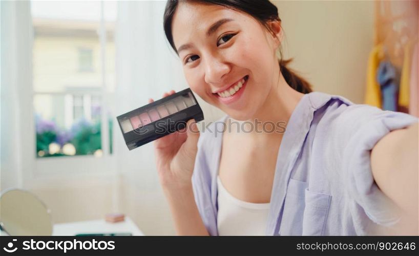 Happy beautiful young Asian woman using smartphone selfie live streaming online to social network. Beauty blogger present beauty cosmetics sitting in front tablet camera for recording video.