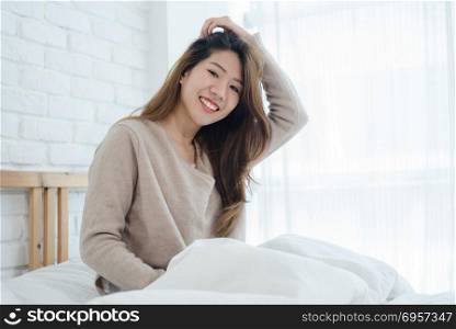 Happy beautiful young Asian woman waking up in morning, sitting . Happy beautiful young Asian woman waking up in morning, sitting on bed, stretching in cozy bedroom, looking through window. Funny asian woman after wake up. Asia woman is stretching and smiling.