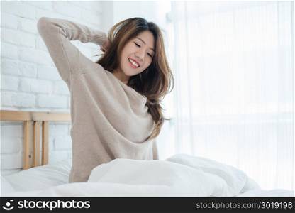 Happy beautiful young Asian woman waking up in morning, sitting on bed, stretching in cozy bedroom, looking through window. Funny asian woman after wake up. Asia woman is stretching and smiling.