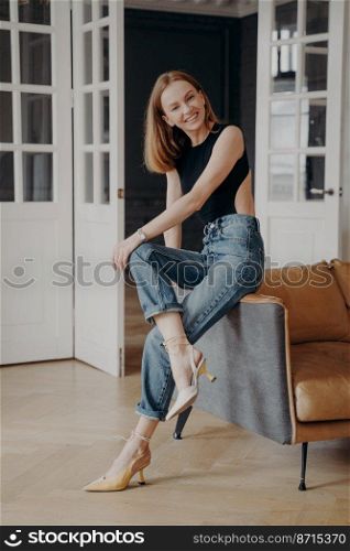 Happy beautiful young female sitting on comfortable sofa looking at camera, posing for portrait indoors. Pretty girl fashion model sits on cozy couch against white doors background at home.. Happy beautiful young girl sitting on comfortable sofa looking at camera posing for portrait at home