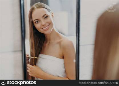 Happy beautiful young woman looks at her reflection in mirror, has well cared long hair, uses hair brush, wrapped in bath towel, makes hairstyle. Women, beauty, hairstyling and self care concept