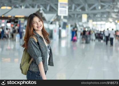 Happy beauty Asian woman traveling and holding suitcase in airport terminal with crowd people passengers background. People and Lifestyle concept. Journey around the world theme. Cheerful tourist girl