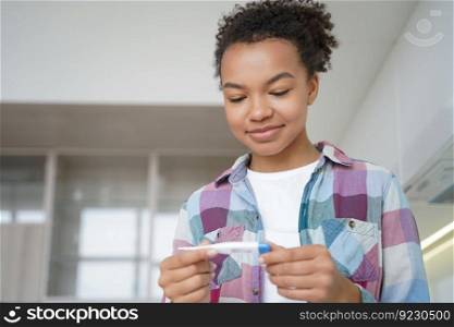 Happy biracial girl holds digital thermometer, satisfied with normal body temperature. Smiling african american teen check temp before trip during covid coronavirus pandemic, pleased with result.. Biracial teen girl holds thermometer, happy with body temperature before trip during covid pandemic