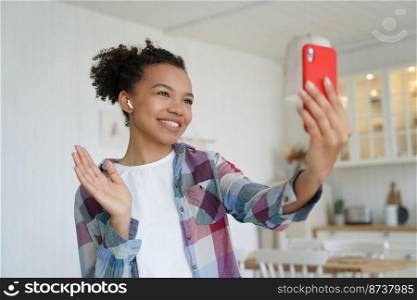 Happy biracial teen girl makes greeting hello gesture chatting online by video call, holding smartphone. Modern young mixed race lady blogger welcomes followers in social network, talking live at home. Smiling mixed race girl makes greeting hello gesture, talking by video call, holding phone at home