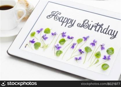 Happy Birthday greeting card - handwriting with viola flowers and leaves on digital tablet with a cup of coffee