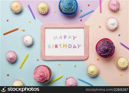happy birthday written wooden frame surrounded with muffins aalaw sprinkles candles colored backdrop