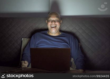 Happy black senior, African American man person watching a funny movie online and laughing on bed in bedroom at home at late night. Activity lifestyle. Relax time. Computer notebook laptop.
