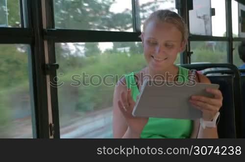Happy blond woman with tablet PC riding in the bus. She texting message on pad sitting by the window with city and traffic view