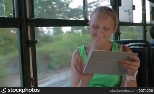 Happy blond woman with tablet PC riding in the bus. She texting message on pad sitting by the window with city and traffic view