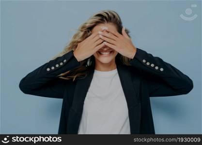 Happy blonde female covering her eyes with both hands and waiting to receive nice gift from boyfriend, excited young woman waiting for pleasant surprise while posing isolated on blue background. Happy blonde female covering her eyes with both hands and waiting to receive nice gift