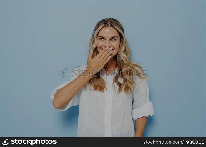 Happy blonde woman closing mouth with hand going to see surprise prepared by friends, excited female smiling in anticipation for something wonderful, studio shot of amazed lady covering face with hand. Young excited female smiling in anticipation for something wonderful