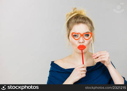 Happy blonde woman holding carnival accessories on stick fake red lips and paper heart shaped glasses, having fun. On grey wall background. woman holds carnival accessories on stick