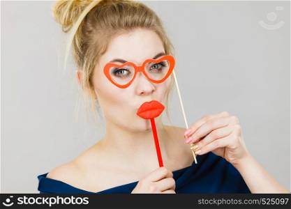 Happy blonde woman holding carnival accessories on stick fake red lips and paper heart shaped glasses, having fun. On grey wall background. woman holds carnival accessories on stick