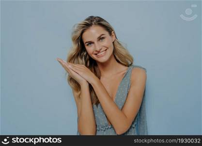 Happy blonde woman wearing blue dress holding hands together next near her head with smile, expressing happiness and positive emotions, standing isolated next to blue background in studio. Happy blonde woman holding hands together next to her head with smile