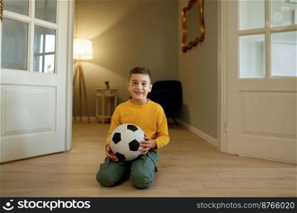 Happy boy football fan posing for camera with soccer ball at home. Little child having fun in living room in evening. Happy boy football fan posing for camera with soccer ball at home