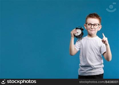 Happy boy in a white t-shirt thinking and looking up with a alarm clock pointing his finger up on a blue background.. Happy boy in a white t-shirt thinking and looking up with a alarm clock pointing his finger up on a blue background