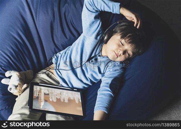 Happy boy lying on bean bag closing his listening music from digital tablet, Positive boy staying at home relaxing after online homework lesson from school during quarantine.Distance education, E-learning and Self Distancing