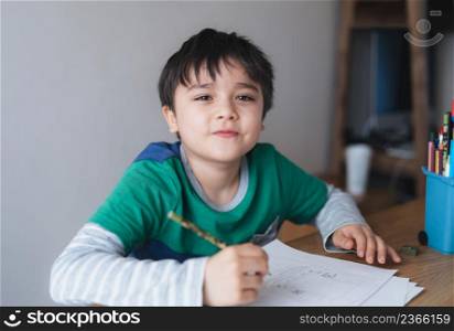 Happy boy using pencil drawing or sketching on paper, Cute kid looking at camera with smiling face siting on table doing homework, Child enjoy art and craft activity at home, Education concept