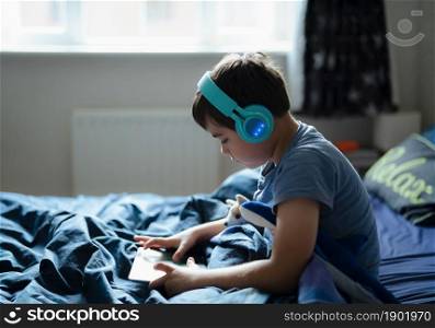 Happy boy wear wireless headphone listening music while sitting in bed, Cute young kid playing game on tablet,Positive child in blue pajamas relaxing in bed room in morning before go to school