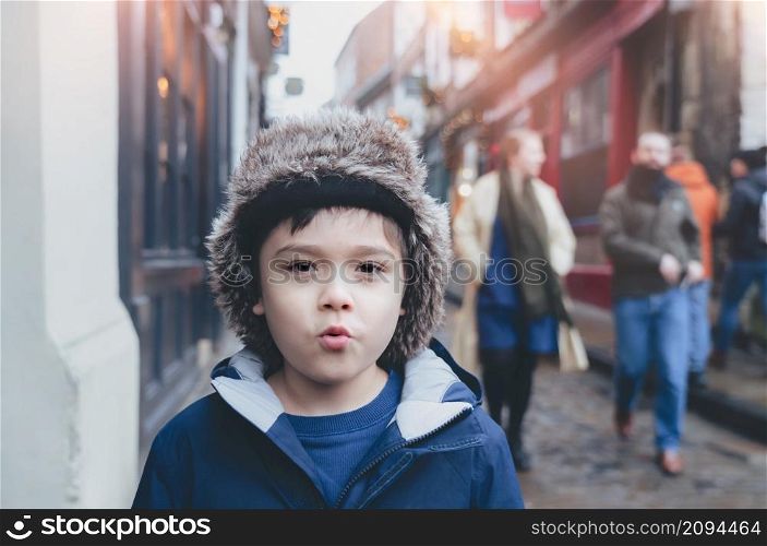 Happy boy with fluffy hat and warm jacket walking outside in the city, Cute kid with funny face standing outside with blurry street background, Child having fun playing outdoor in old town on Christmas holiday.