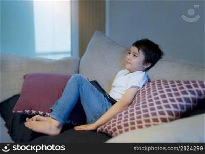 Happy boy with smiling face sitting on sofa watching cartoon, School Kid sitting alone on couch holding tablet and watching TV, Portrait Child relaxing in living room after back from school.