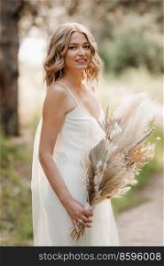 happy bride girl in a white light dress with a bouquet of dried flowers on a forest path
