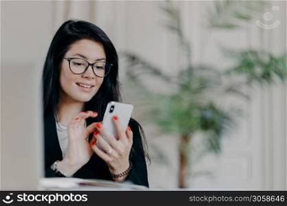 Happy brunette businesswoman uses smartphone, wears spectacles, sends text messages to partner, works on project, poses against cozy interior at workplace. Technology, occupation and work concept