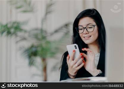 Happy brunette businesswoman uses smartphone, wears spectacles, sends text messages to partner, works on project, poses against cozy interior at workplace. Technology, occupation and work concept