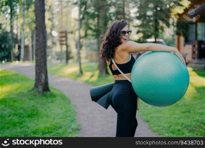 Happy brunette carries fitball and fitness mat in sport clothes, poses in green park. Good physical shape showcases healthy lifestyle and hobby.