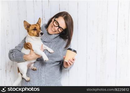 Happy brunette female model wears warm knitted sweater, glasses, carries her favourite dog and takeaway coffee, going to have walk together, have good friendly relationships. People and pets concept