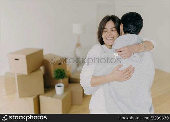 Happy brunette smiling woman embraces with love her husband, holds keys, buy first house together, purchase new property, pose in unfurnished room with pile of cardboard boxes. Ownership concept