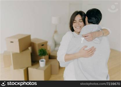 Happy brunette smiling woman embraces with love her husband, holds keys, buy first house together, purchase new property, pose in unfurnished room with pile of cardboard boxes. Ownership concept