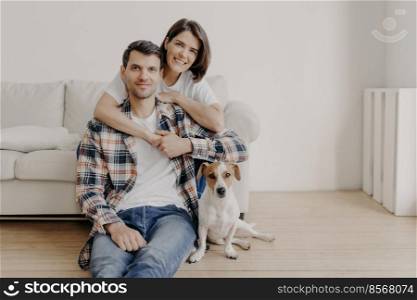 Happy brunette woman embraces husband with love, being in good mood, smiles positively. Husband, wife and dog pose together in living room of new dwelling, enjoy comfort. Couple in love indoor