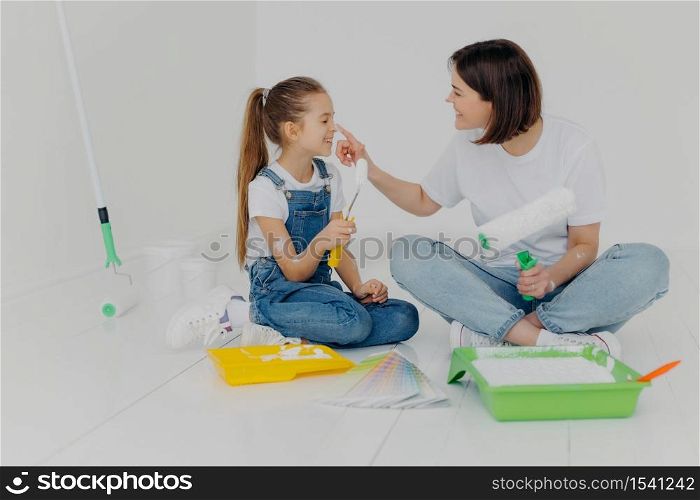 Happy brunette woman plays with daughter, have fun after painting and refurbishment of room, smeark each other with paint, choose color from sample, hold paint rollers, sit on floor in casual wear