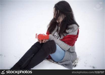 Happy brunette woman with long hair, dressed for cold weather with high boots, sitting in a field of snow while holding a red gift box in her hands.