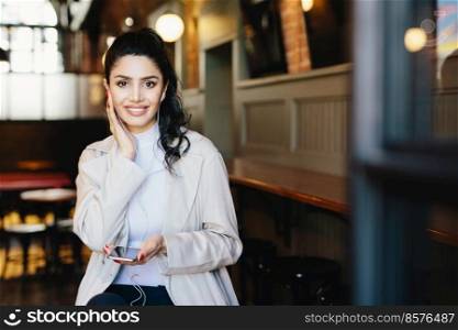 Happy brunette woman with make-up and manicure wearing white jacket holding smartphone and having earphones in ears listening to music or audiobook on earphones. Dreaming female in cafe using mobile