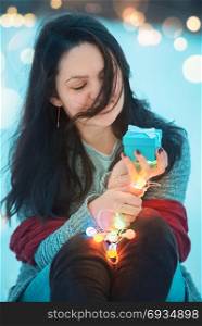 Happy brunette young woman with long hair and dressed for cold weather, holding a blue gift box and colorful lit string lights, outdoor in the snow.