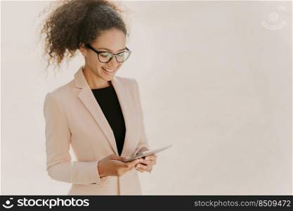 Happy business lady stands with touchpad, reads news online, wears spectacles and elegant clothes, has curly hair combed in pony tail, stands over white wall with blank space, looks for information
