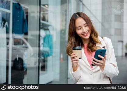Happy business woman hold mobile phone and paper cup of hot drink outdoor walking on city street, Asian businesswoman holding coffee cup takeaway and smartphone go to work she walking near her office