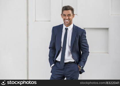 Happy businessman in suit holding hands in pockets and looking at camera with smile against white background on city street in daytime. Cheerful male executive against white wall