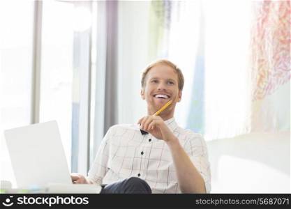 Happy businessman looking away while sitting at desk in creative office