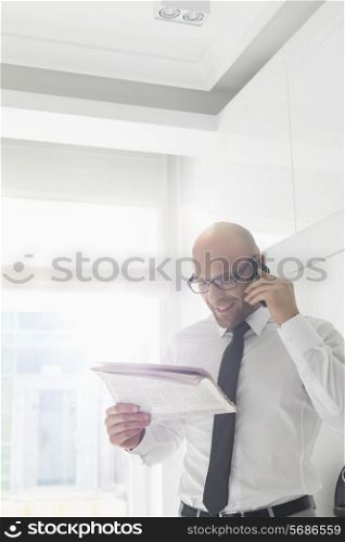 Happy businessman on call while reading newspaper at home