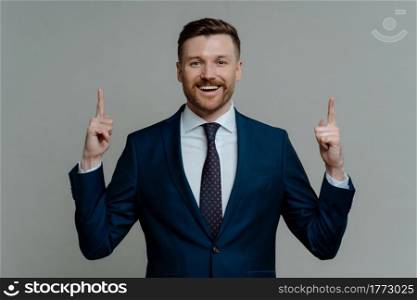 Happy businessman or executive director in suit pointing up with two forefingers and feeling excited while getting business solution or idea, sitisfied entrepreneur standing against grey background. Happy businessman in suit pointing up with fingers and smiling at camera