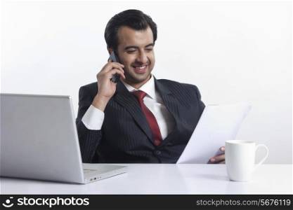 Happy businessman reading document while answering smart phone at desk over white background