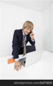 Happy businessman using cell phone while looking at laptop in office