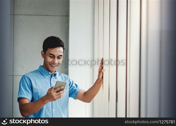 Happy Businessman Using Smartphone. Standing by the Window in Office. Reading Message via Mobile Phone and Smiling